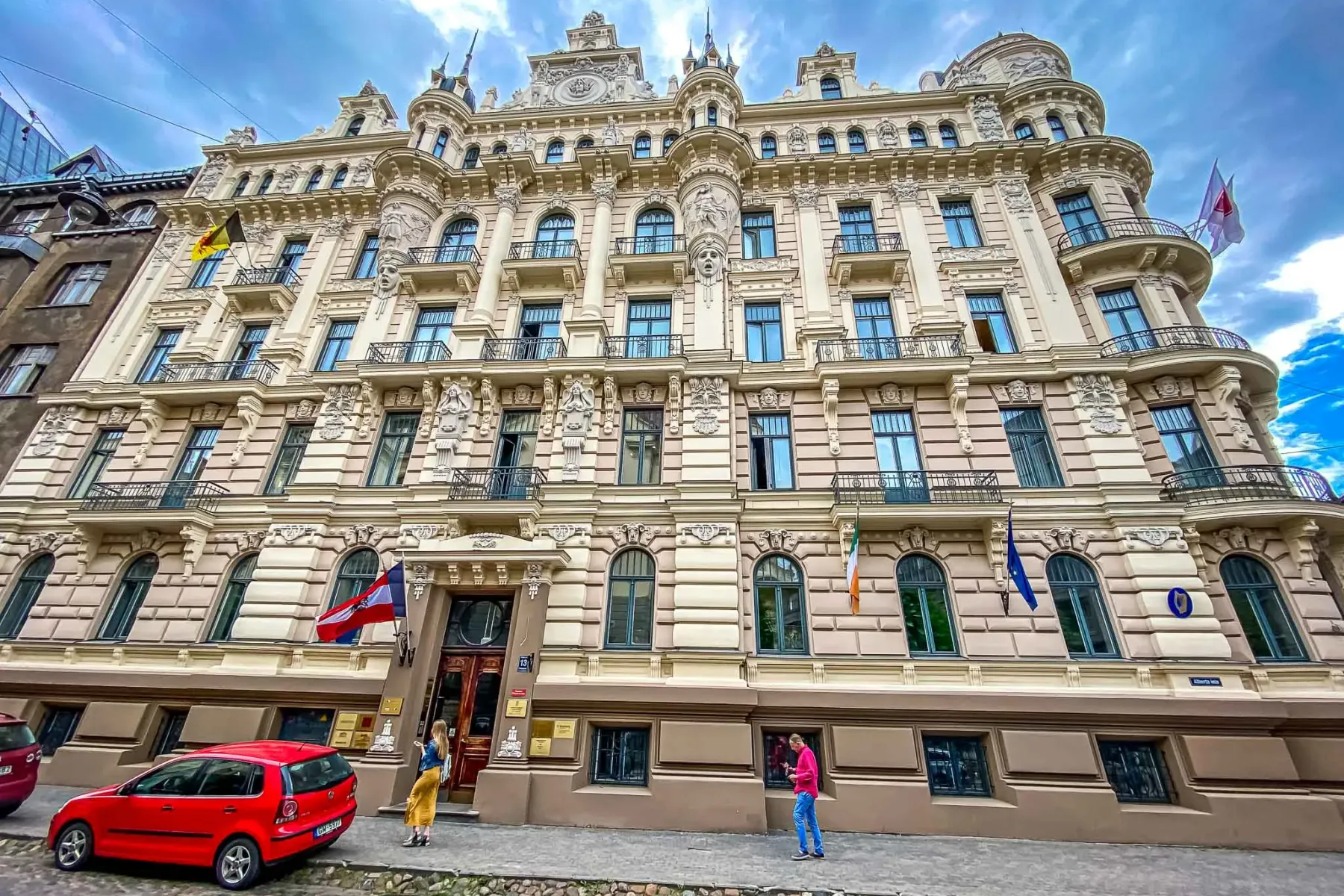 Ornate Art Nouveau building on Alberta Street in Riga, with decorative facade, balconies, and various flags. A red car is ...