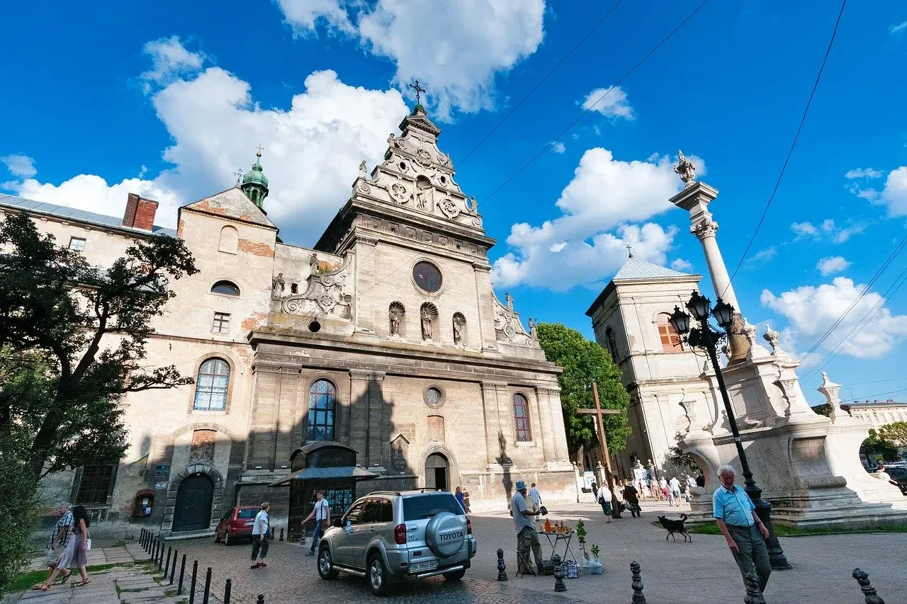 Baroque Church of St. Andrew's in Lviv with a tall ornate Plague Column on a sunny day. People walk along the street.
