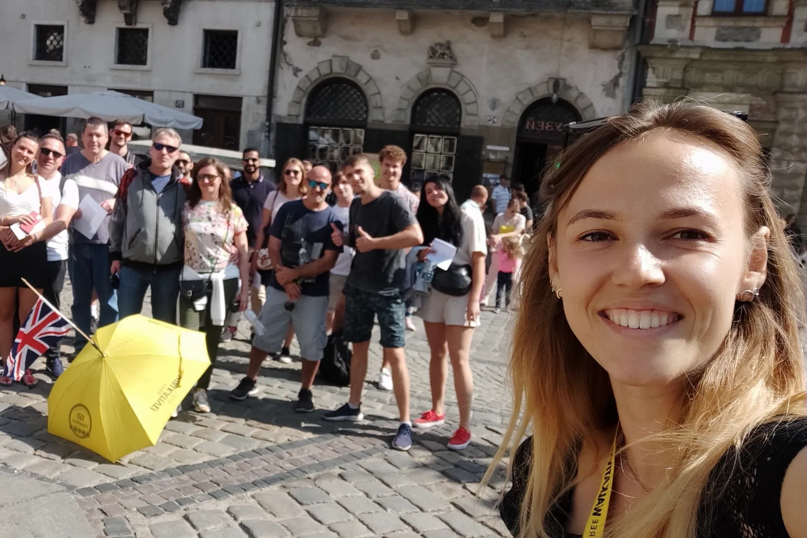 Smiling woman takes a selfie with a group of tourists on a cobblestone street in Lviv. A yellow umbrella and Union Jack fl...