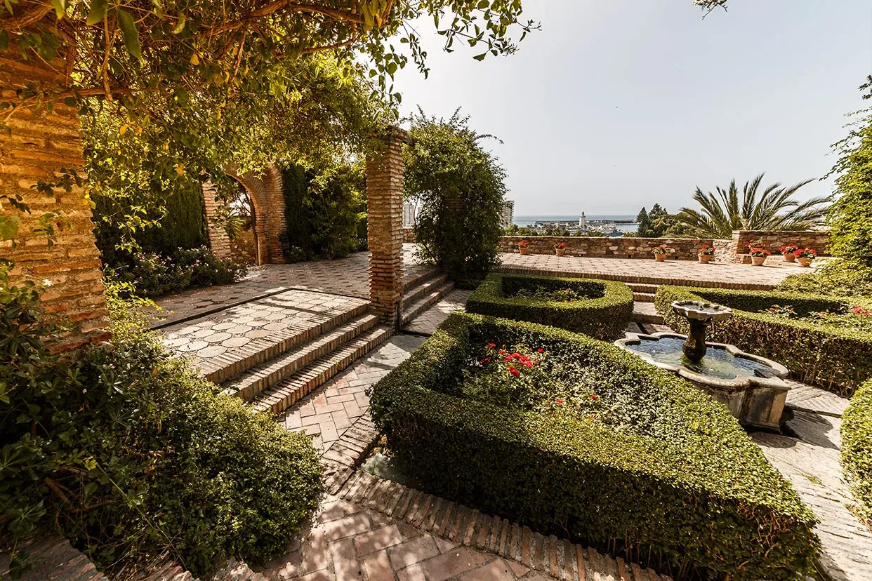 Sunny terraced garden with brick paths, steps, trimmed hedges, a stone fountain, and distant sea view.
