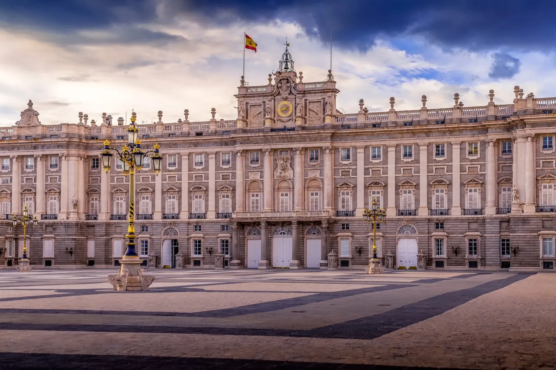 Grand Royal Palace of Madrid with a Spanish flag on top, two ornate street lamps, and a wide empty plaza under a dramatic ...