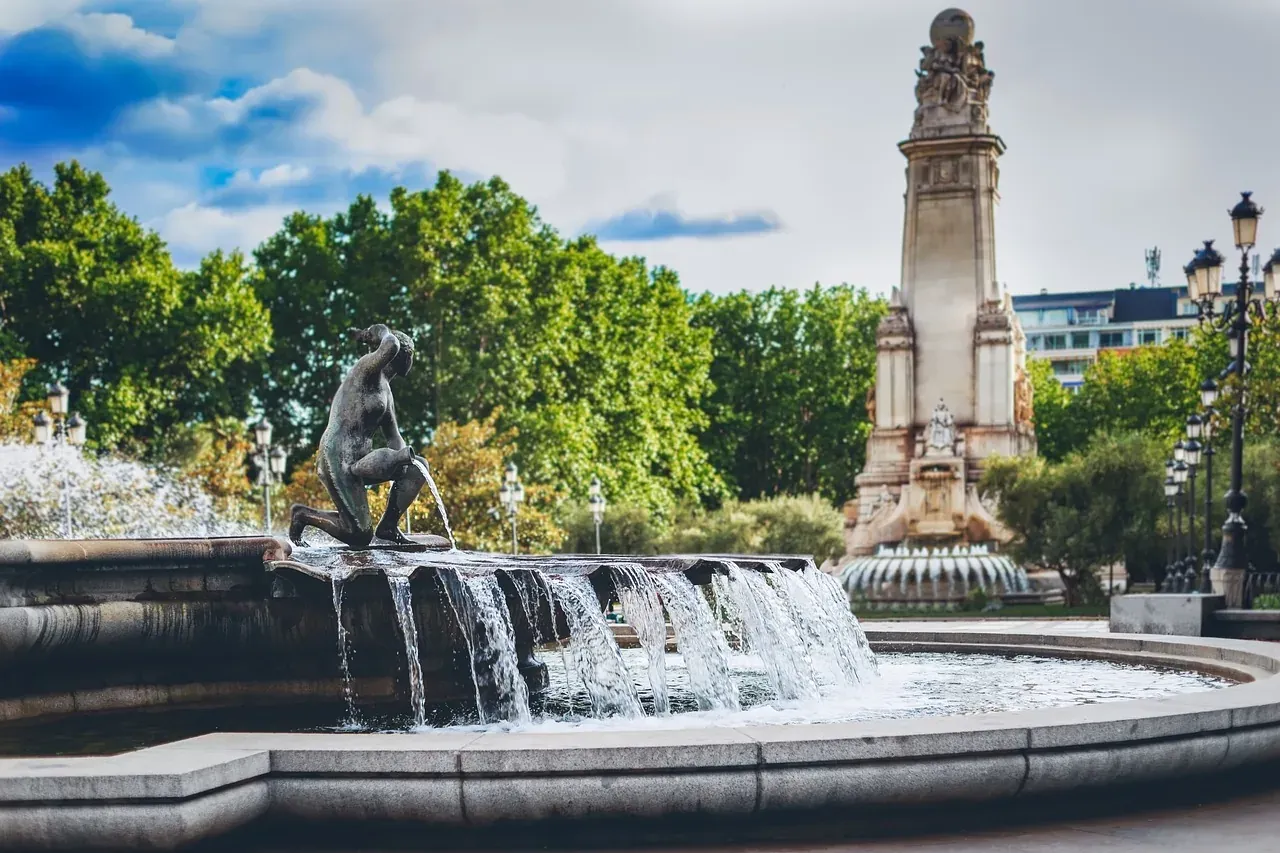 A kneeling bronze sculpture on a tiered fountain, with water flowing. In the background, a tall stone monument and green t...