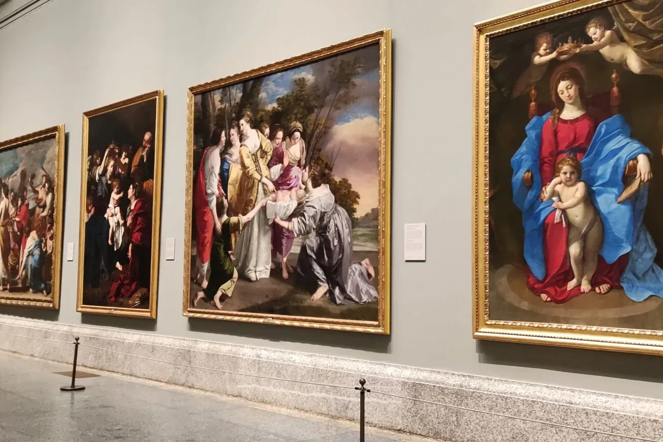 Four large, framed classical paintings hang on a light grey museum wall, with ropes and stanchions on the floor.