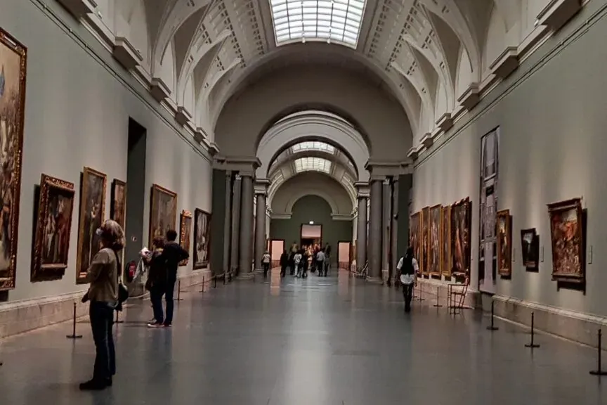 Long, high-ceilinged museum hall with framed paintings on both walls and a large rectangular skylight in the ceiling. Peop...