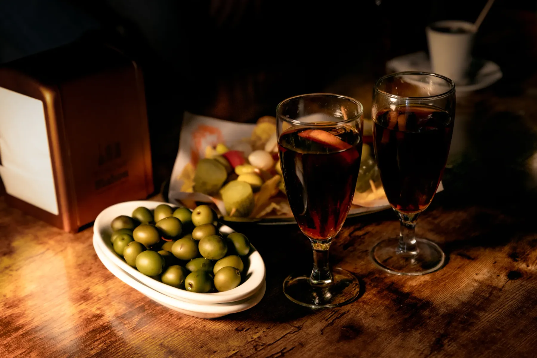 Two glasses of red vermouth and a white bowl of green olives on a wooden table, with tapas and a napkin dispenser in backg...