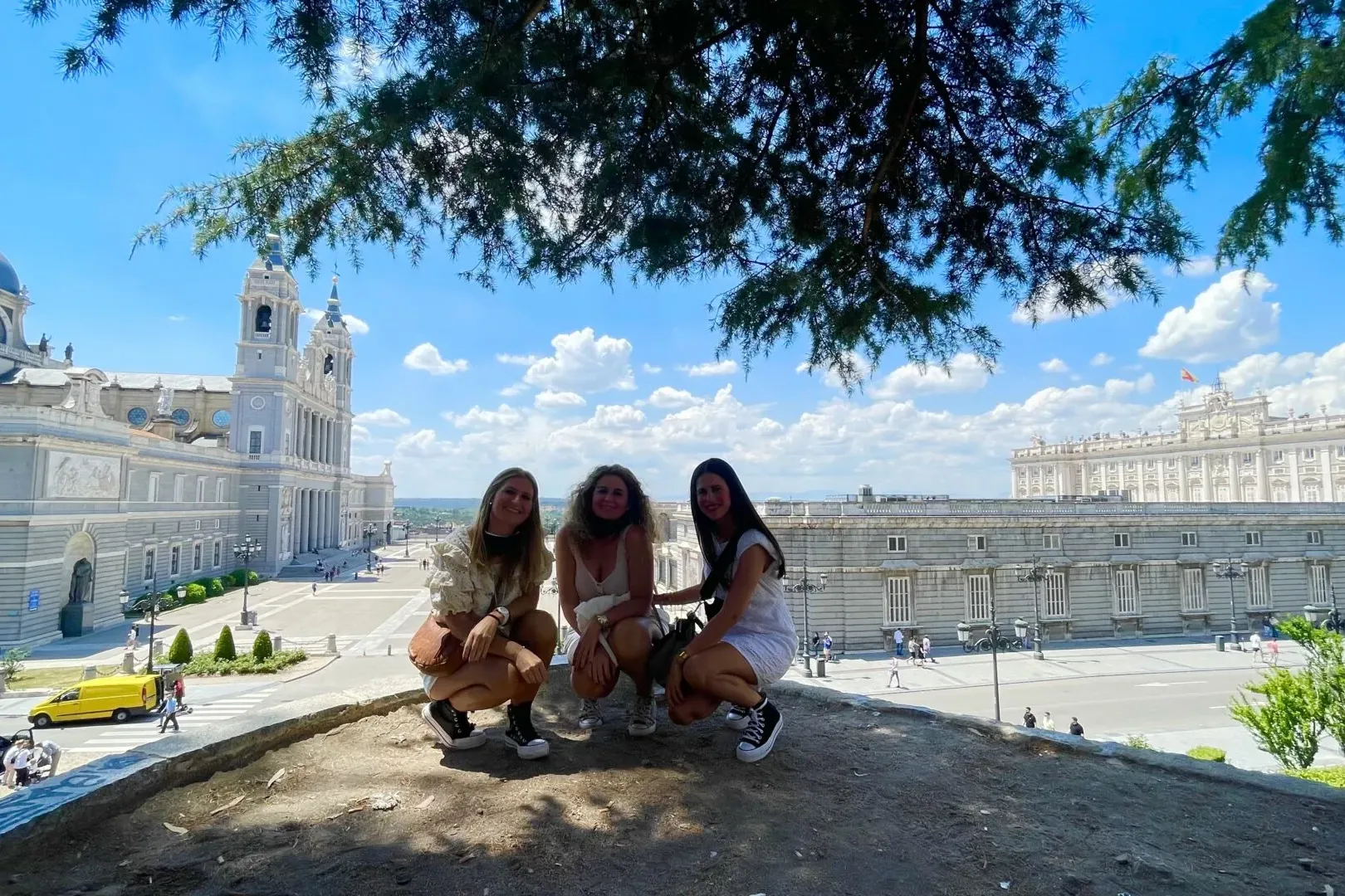 Three women squatting in front of Almudena Cathedral and the Royal Palace of Madrid under a blue sky.