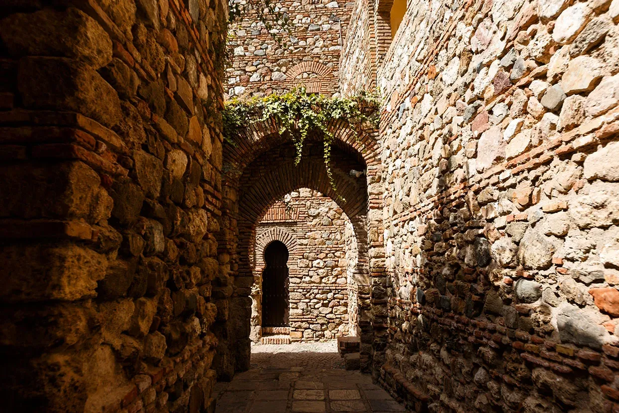 Narrow stone alley with high walls, multiple brick arches, and a dark wooden door with an arched top. Green ivy on the upp...