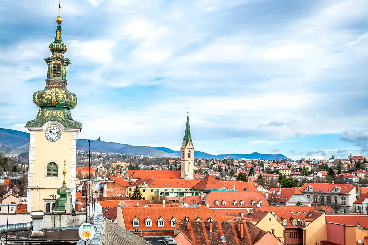 View of Zagreb's Old Town featuring a baroque clock tower, many red-tiled roofs, and distant hills under a cloudy sky.