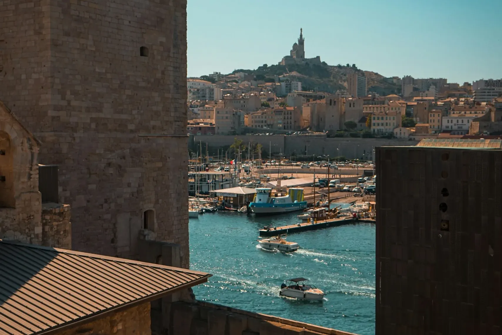 View from a stone fortress over Marseille's Old Port with boats and Notre-Dame de la Garde basilica on a hill.