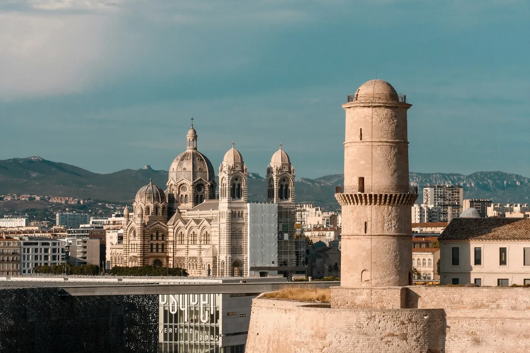 Marseille's Fort Saint-Jean tower and walls by the sea, with Cathédrale La Major, Mucem museum, and distant mountains.