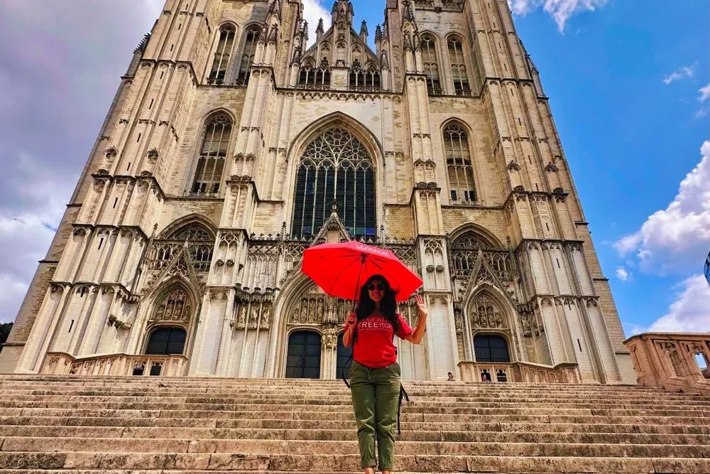 A woman holding a red umbrella stands on wide stone steps leading up to the grand Gothic Cathedral of St. Michael and St. ...