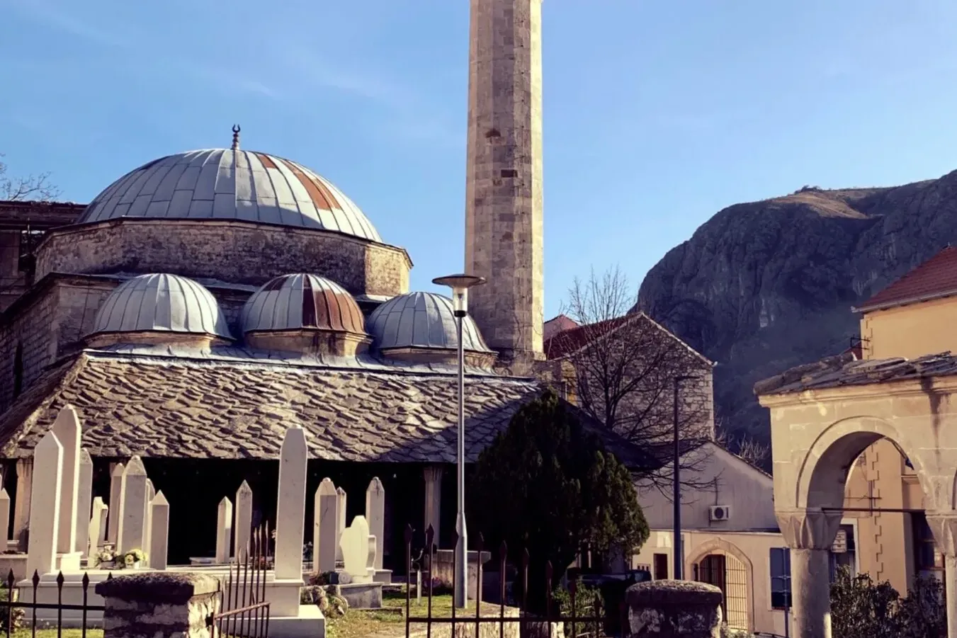 A stone mosque with multiple domes and a tall minaret, surrounded by a graveyard with white tombstones under a blue sky.