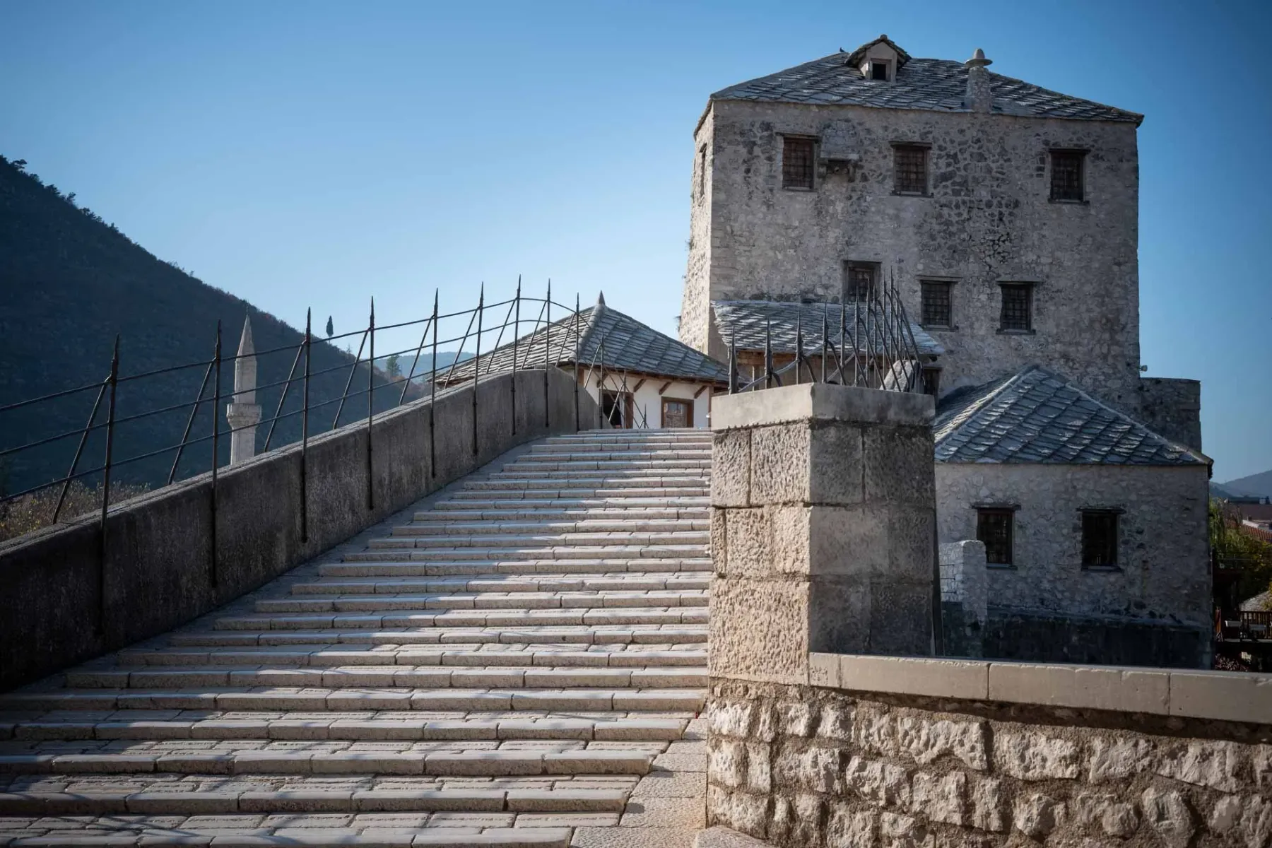 Stone steps of an old bridge leading up to a fortified stone building with a slate roof. A minaret and mountains are in th...