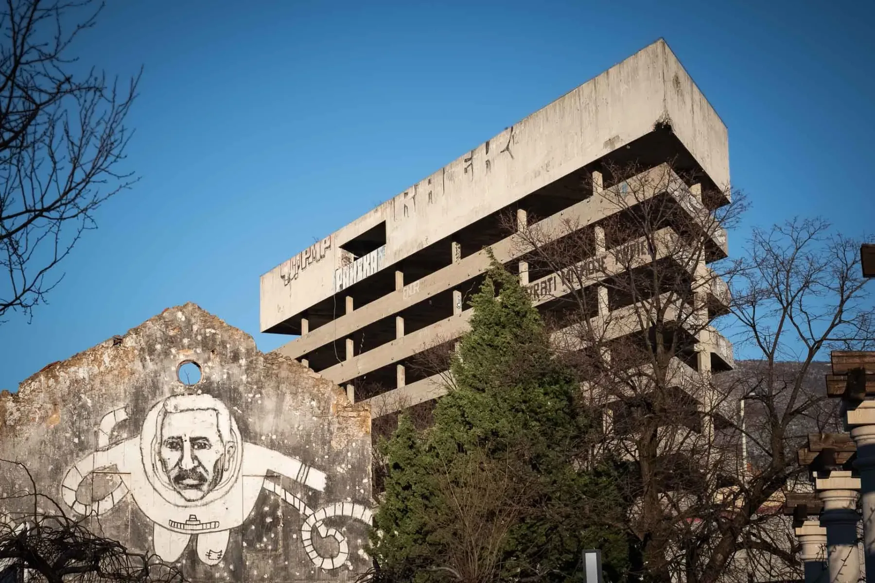 Dilapidated concrete building with graffiti and a ruined wall featuring a large mural of a man in a spacesuit, Mostar.