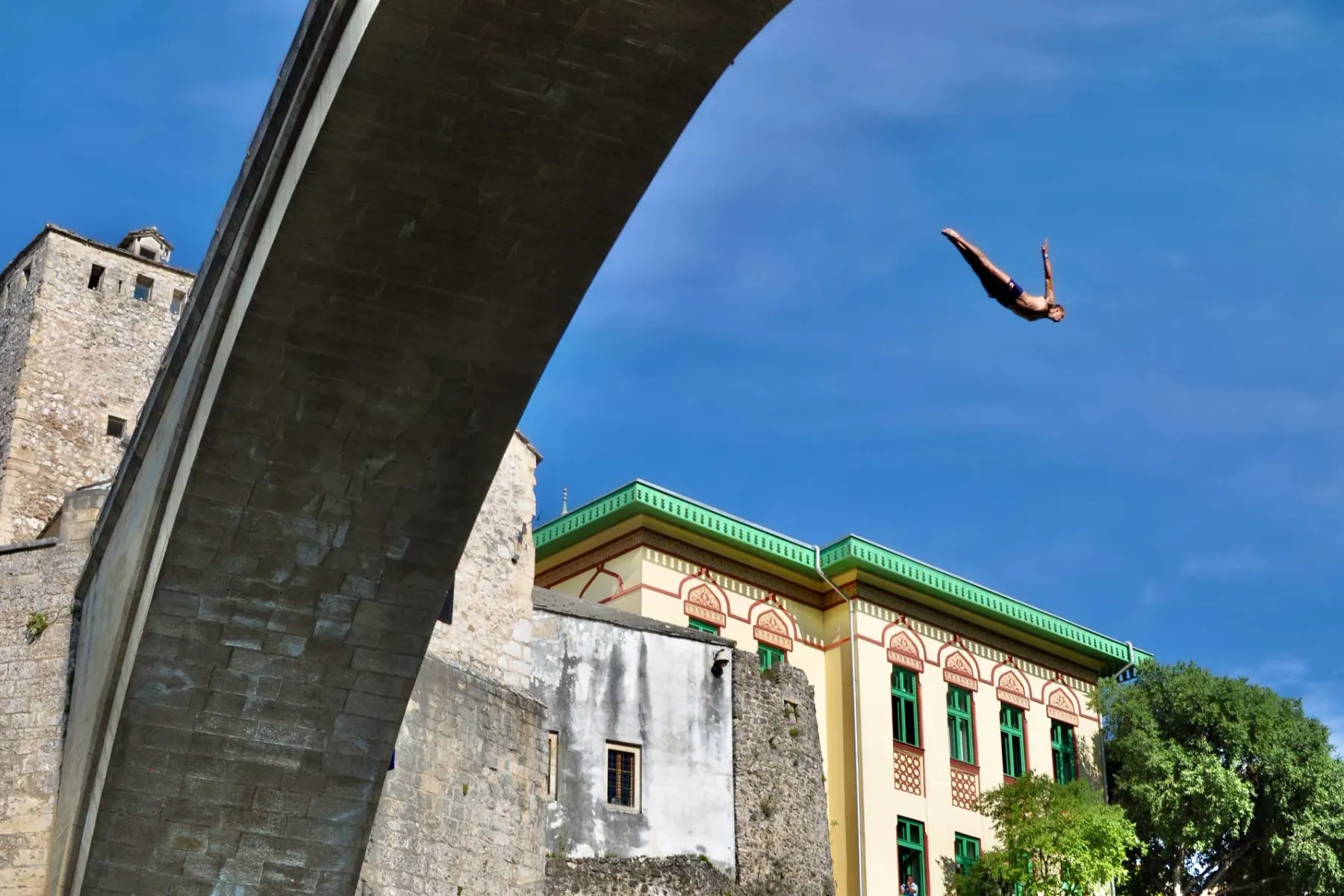 A man in blue swim shorts dives from Stari Most bridge against a clear blue sky, with traditional buildings visible below.