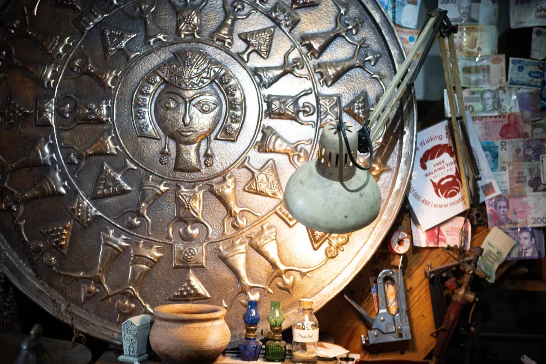 Large circular embossed metal plate with a stylized face, surrounded by patterns. A white desk lamp illuminates it, with c...