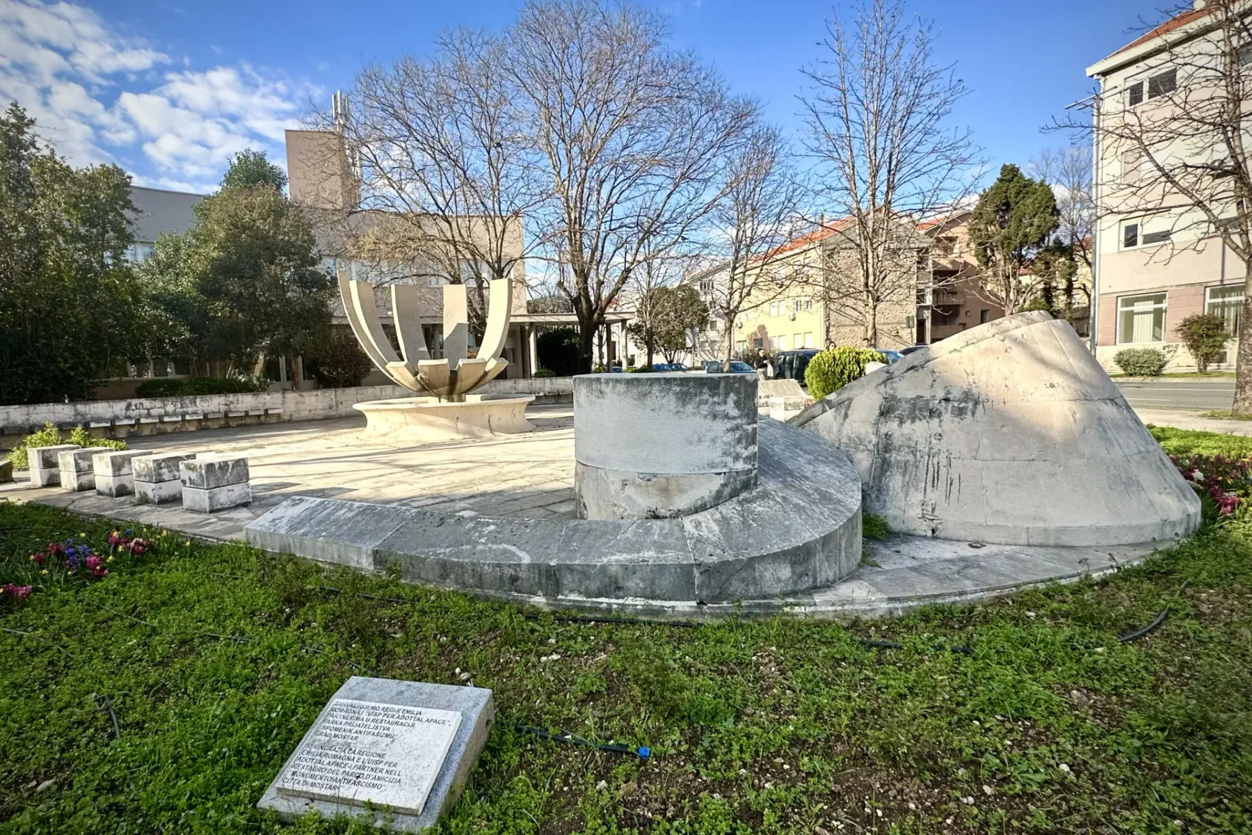 Concrete anti-fascist monument with abstract shapes in Mostar, a square with bare trees and buildings under a blue sky.