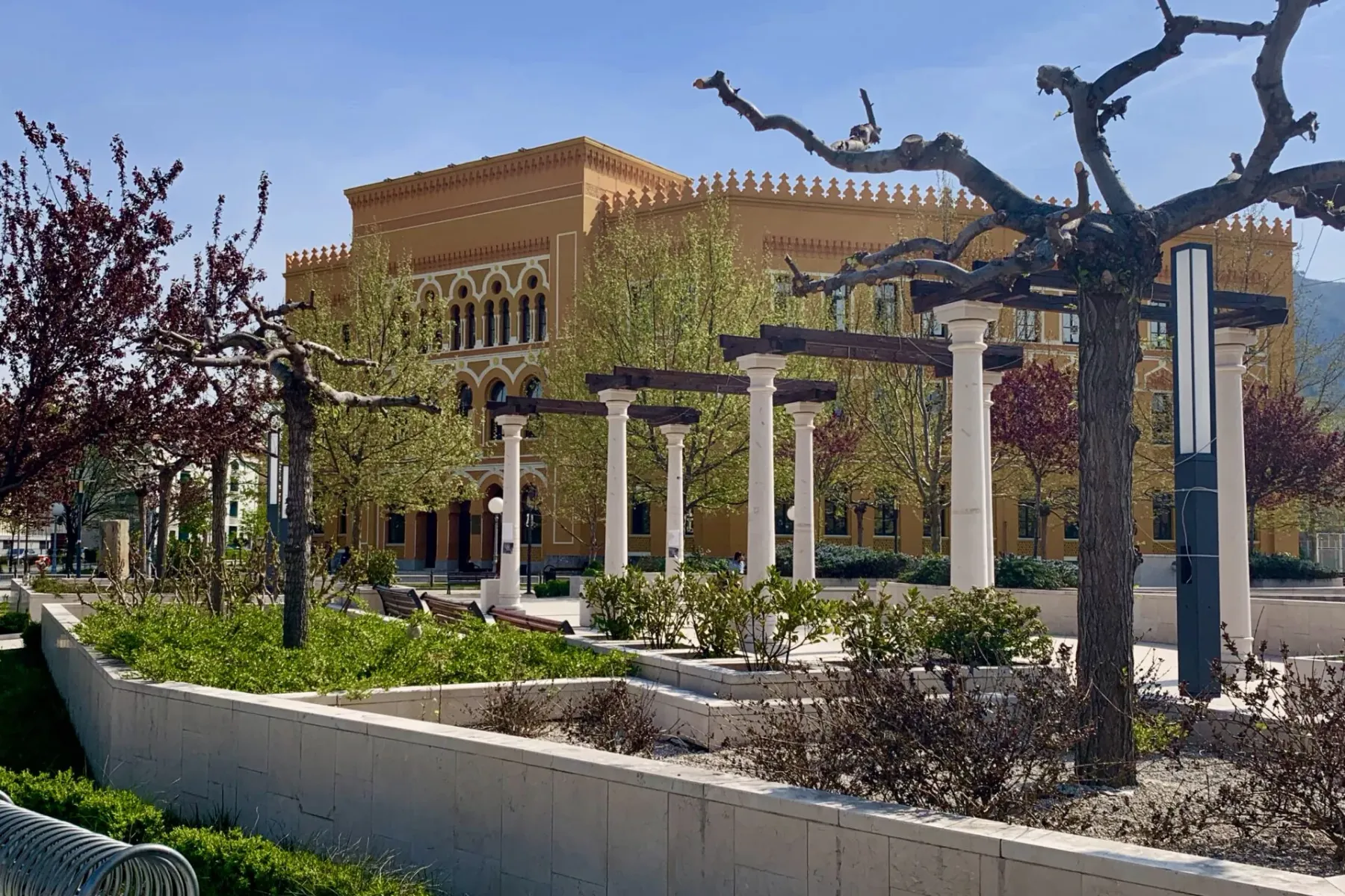 A grand yellow building with arched windows behind a park with white columns and leafless trees under a clear blue sky.