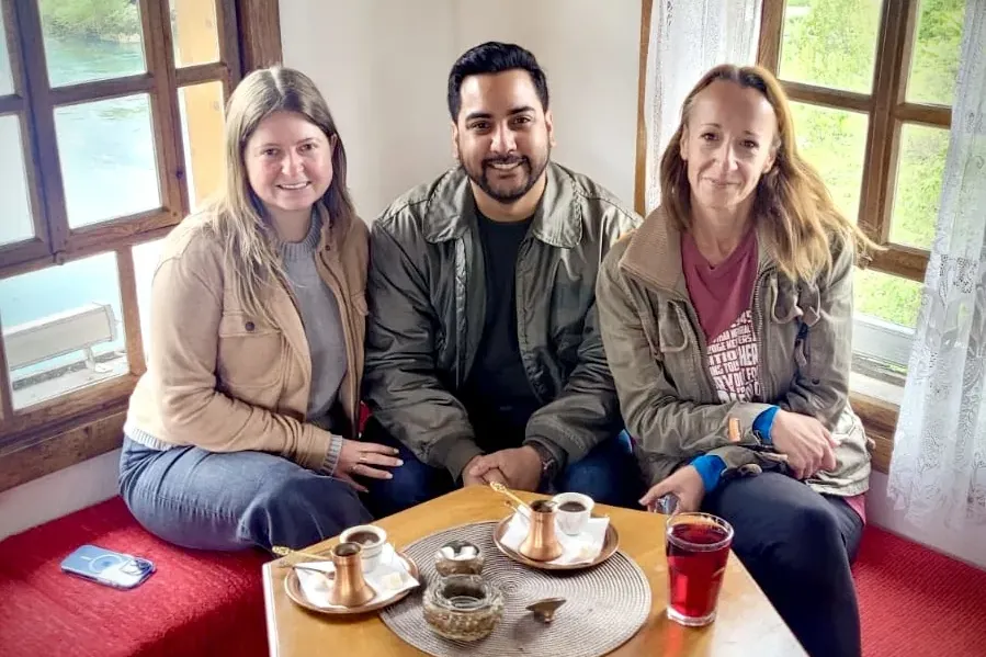 Three travellers enjoying traditional Bosnian coffee and a drink at a wooden table by a window overlooking a river.
