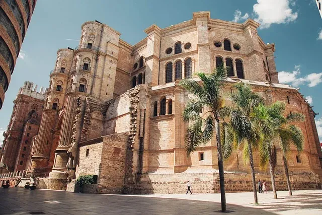 Large stone cathedral with ornate baroque and renaissance details, one main tower, palm trees, and people on a sunny plaza.