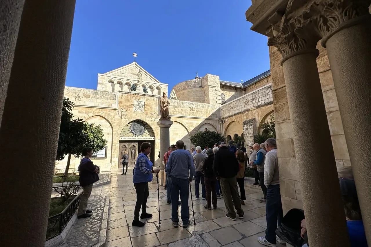 Tourists in a stone courtyard, facing a historic church facade with arches, a statue, and a cross on top, under blue sky.