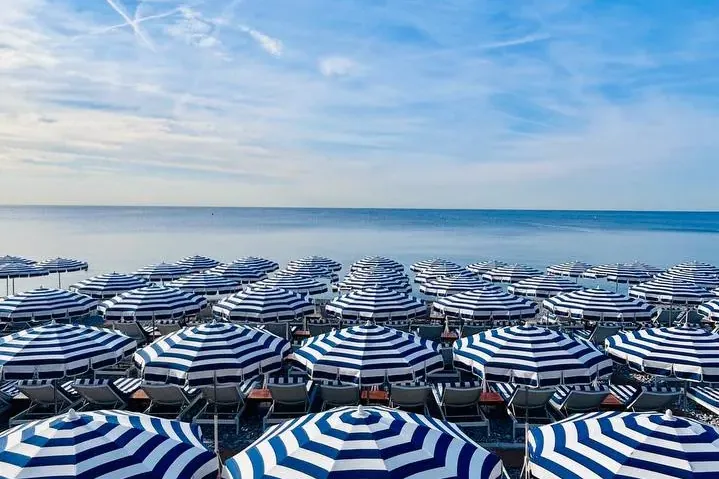 Rows of blue and white striped beach umbrellas and lounge chairs on a pebble beach, facing the calm sea under a clear blue...