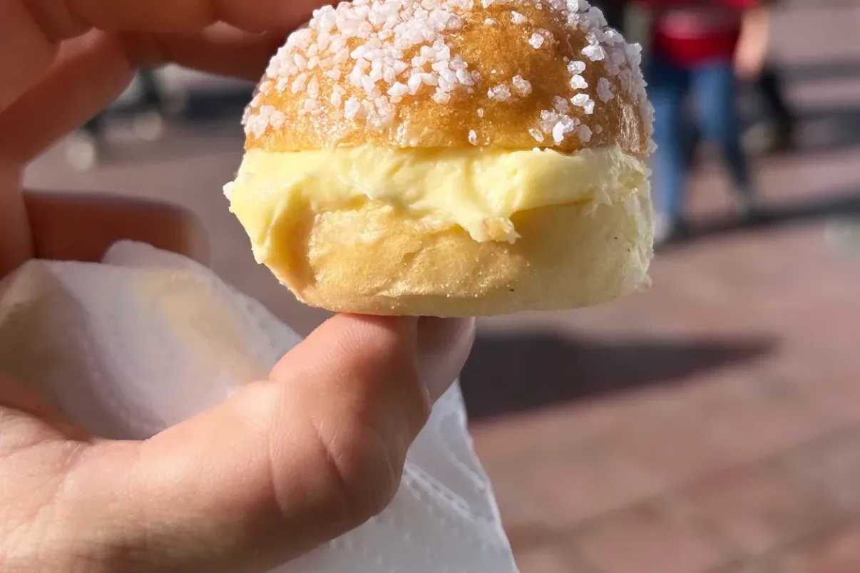 Hand holding a small brioche-like pastry with cream filling and coarse sugar topping. People and pavement are blurred in t...