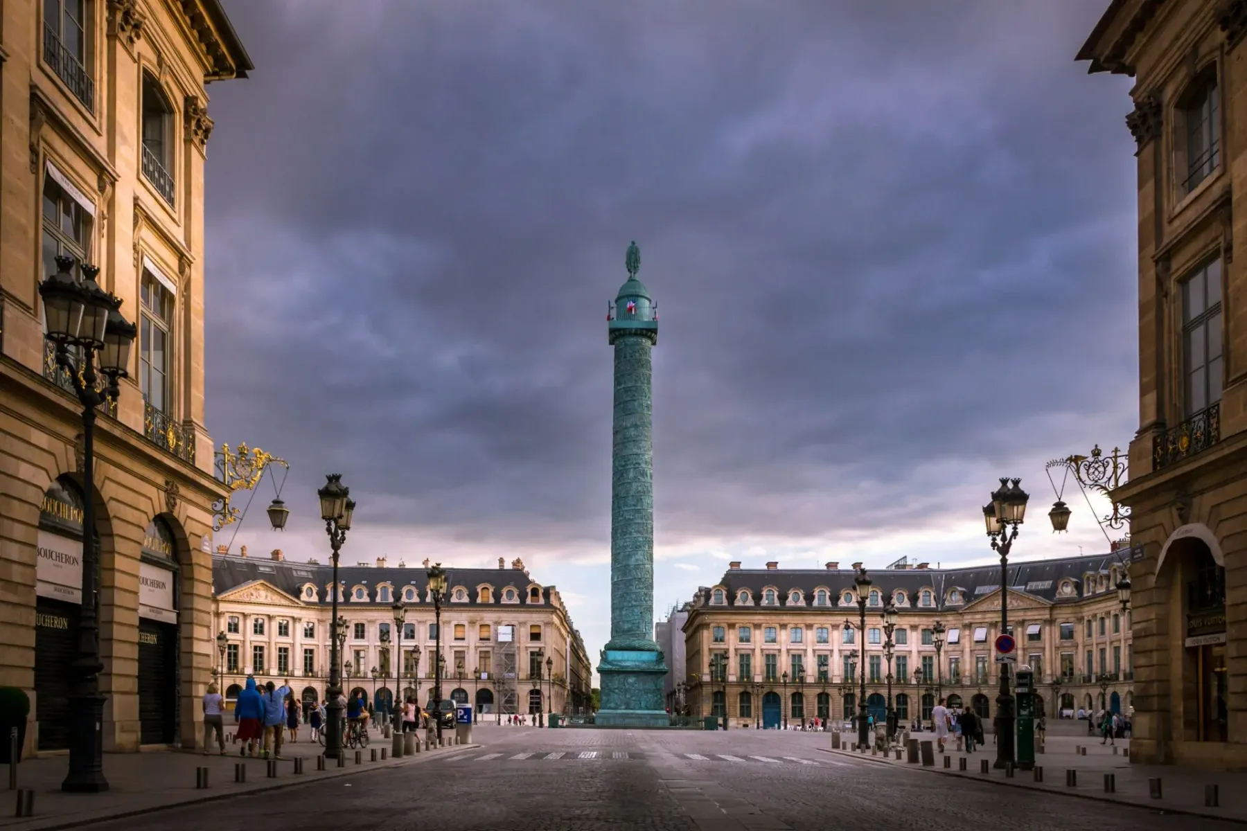 The Vendôme Column in Place Vendôme, Paris, framed by elegant buildings under a cloudy sky. Pedestrians walk on cobbled st...