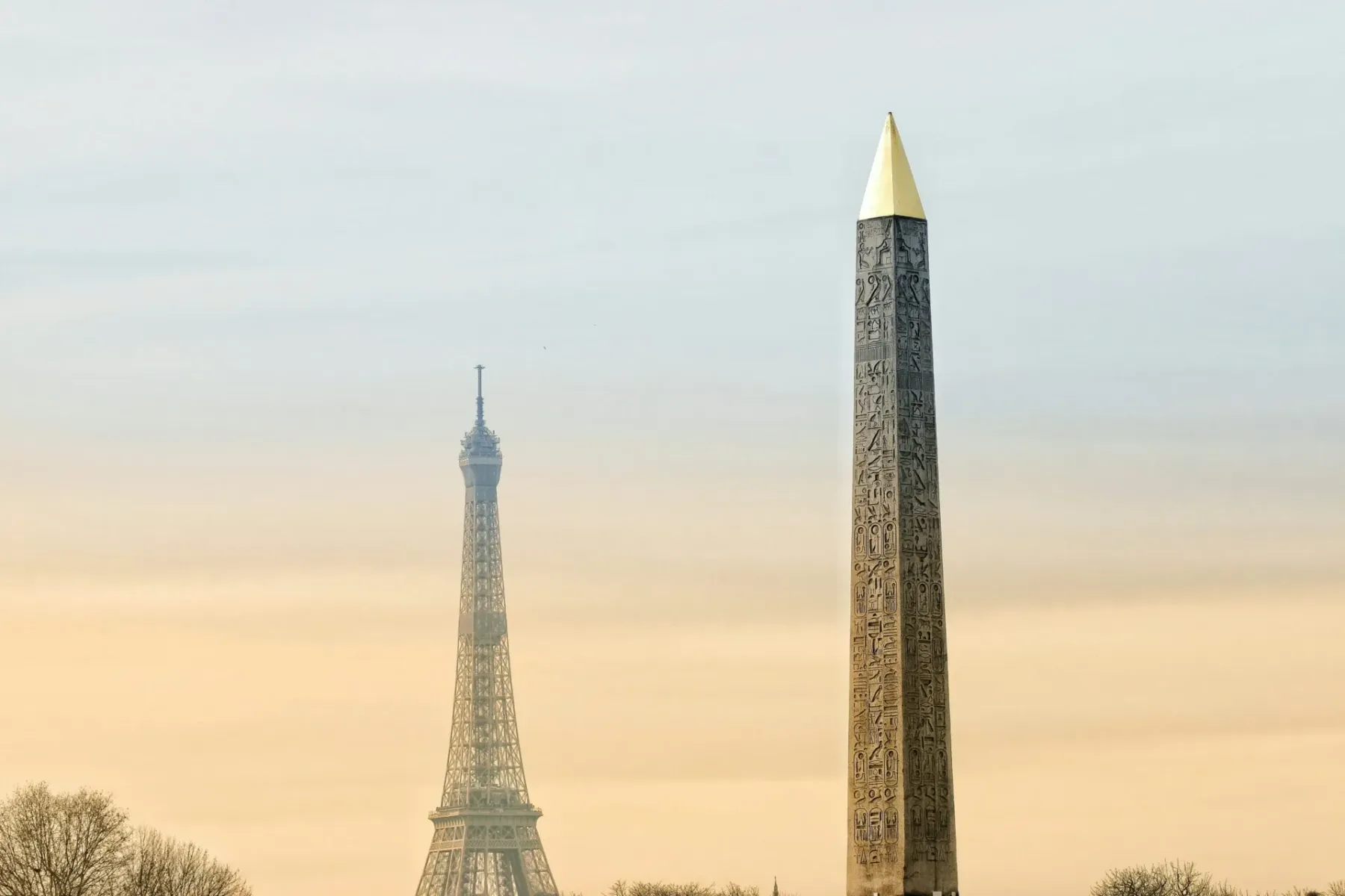 The Luxor Obelisk with a gold cap and ancient hieroglyphs, with the top of the Eiffel Tower visible in the hazy background.