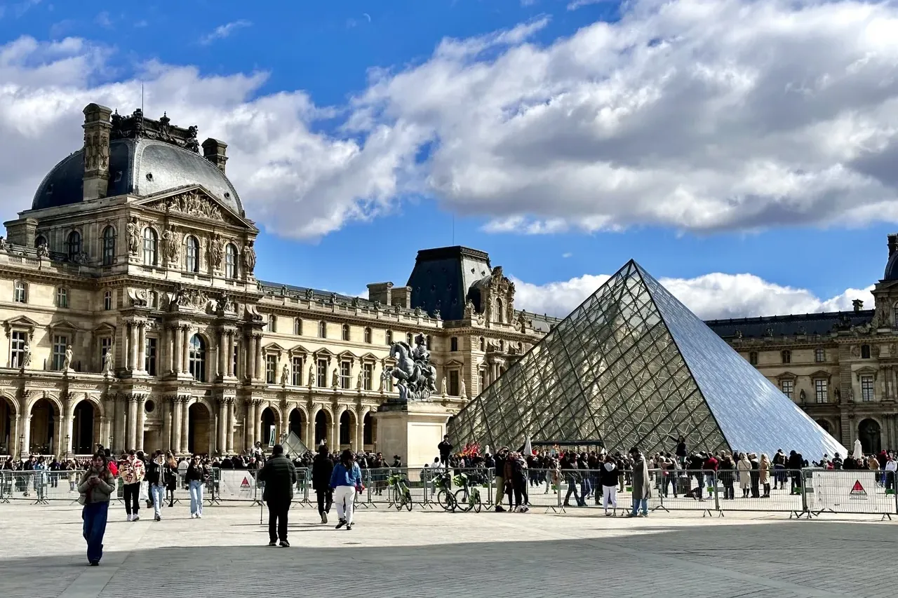 View of the Louvre Museum with its classical architecture and glass pyramid, under a partly cloudy blue sky. People are vi...