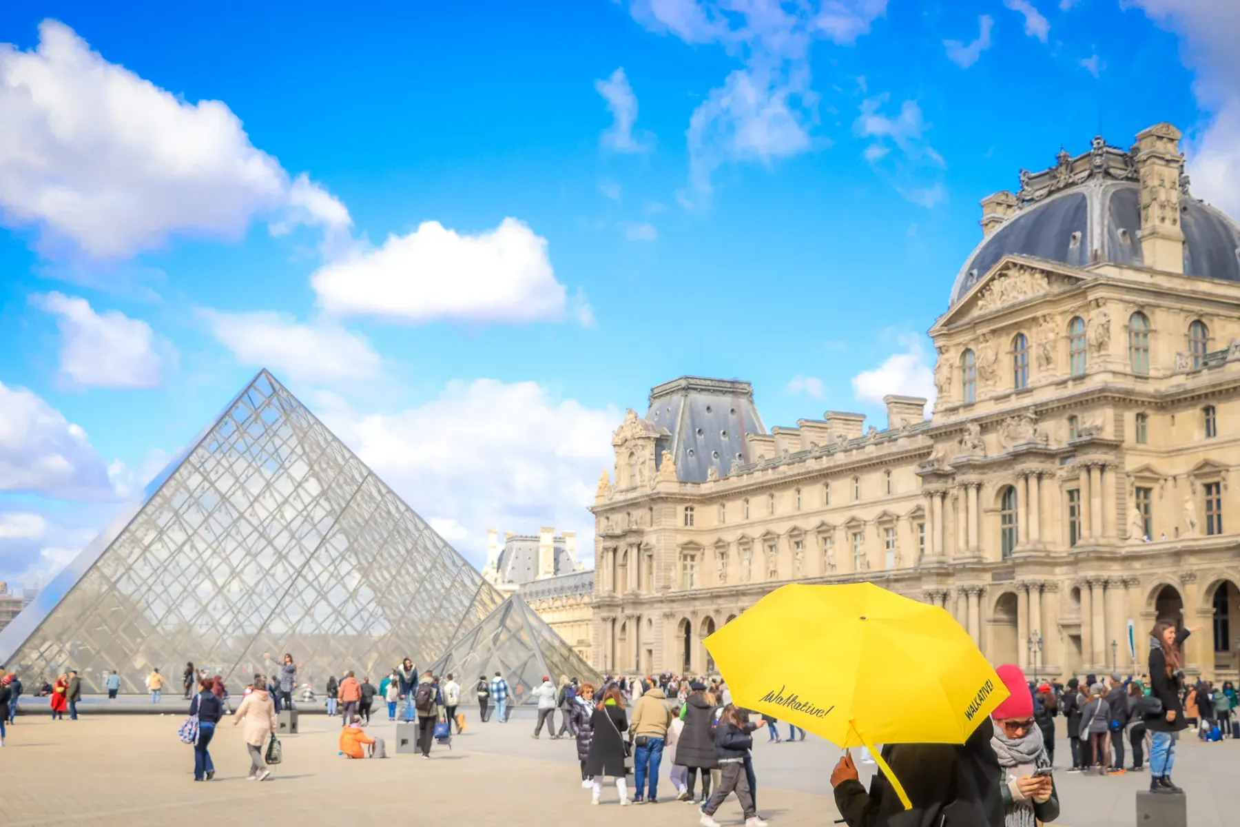 The glass pyramid entrance of the Louvre Museum on a sunny day with clouds, surrounded by classical palace buildings and v...