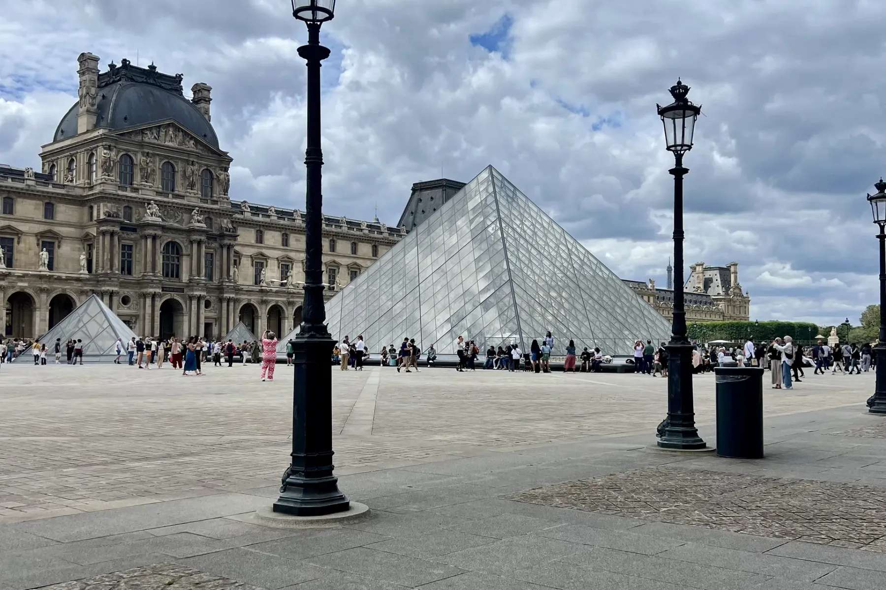 The glass pyramid of the Louvre Museum in Paris, with the historic palace building behind it and people walking on the plaza.