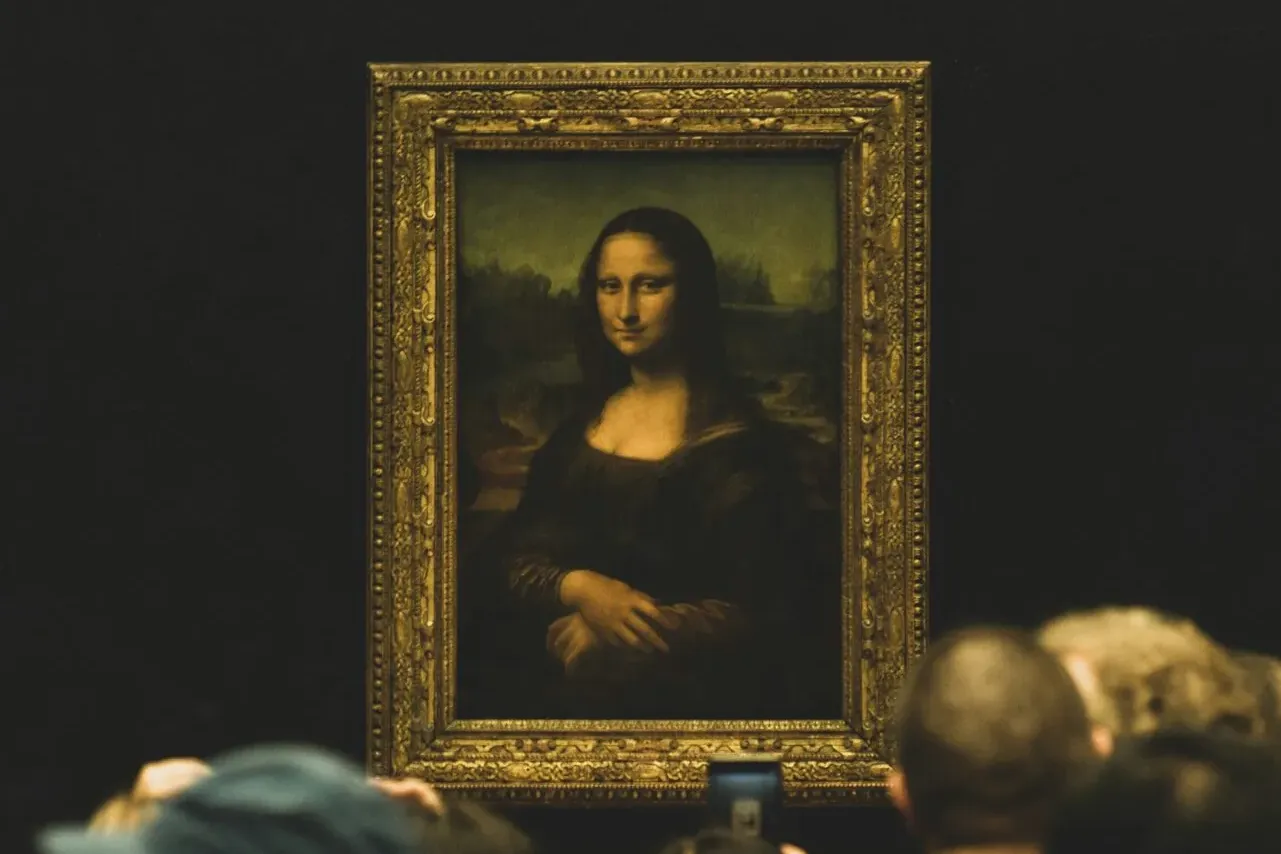 The Mona Lisa painting in an ornate frame, displayed on a dark wall with blurred visitors in the foreground.