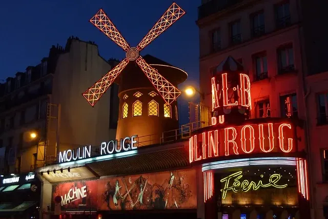 The Moulin Rouge cabaret building at night, illuminated with vibrant red neon signs and a glowing red windmill on the roof.