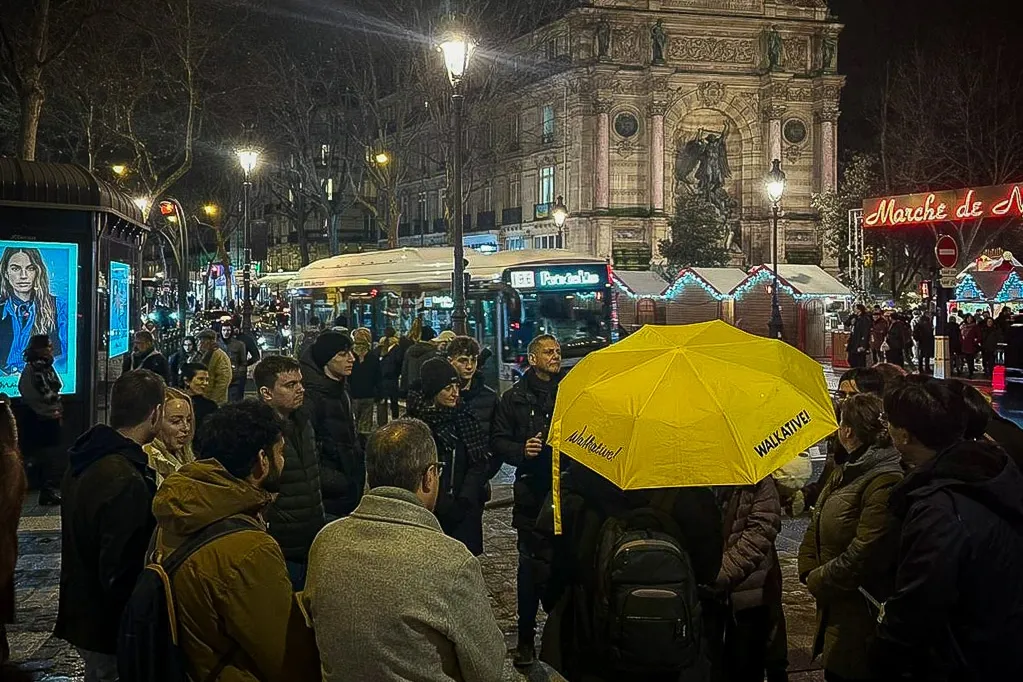 Walkative tour group at night, gathered near the illuminated Fontaine Molière in Paris, with Christmas market stalls visible.