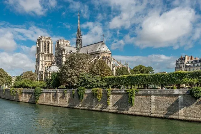 Notre Dame Cathedral in Paris, seen from the Seine River, with its Gothic spires and towers under a partly cloudy sky.