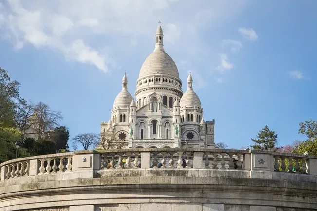 The Sacré-Cœur Basilica in Paris, a white domed church, viewed from below a curved stone balustrade under a blue sky.