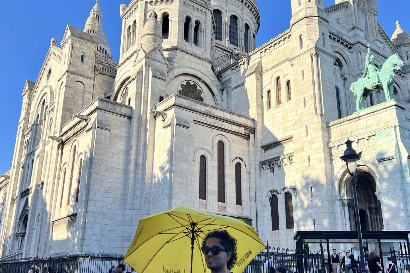Sacré-Cœur Basilica's white domes and facade under a clear blue sky. A person with a yellow umbrella stands on cobblestones.