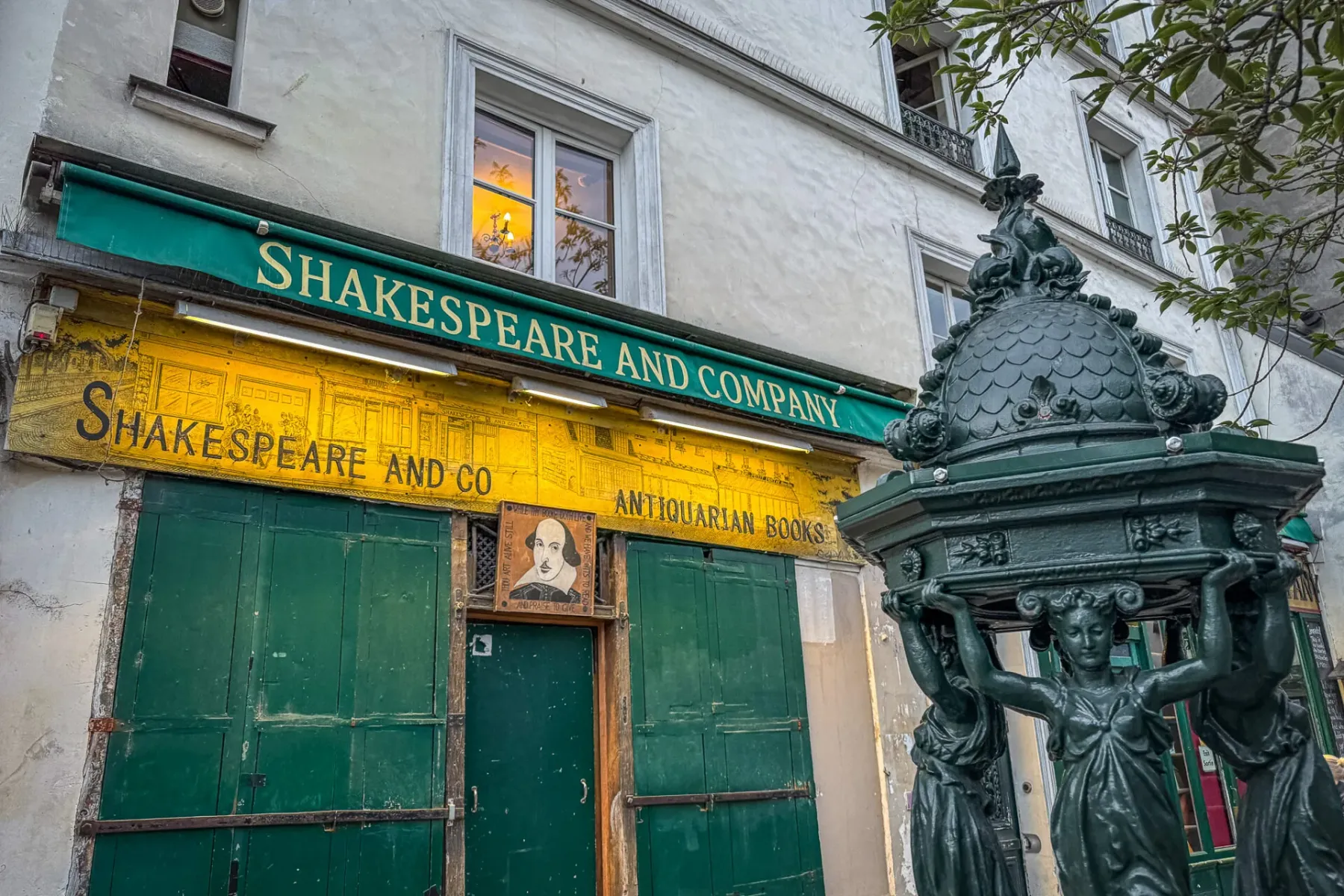 Exterior of Shakespeare and Company bookstore in Paris, with a green awning, yellow facade, and a dark green Wallace fount...