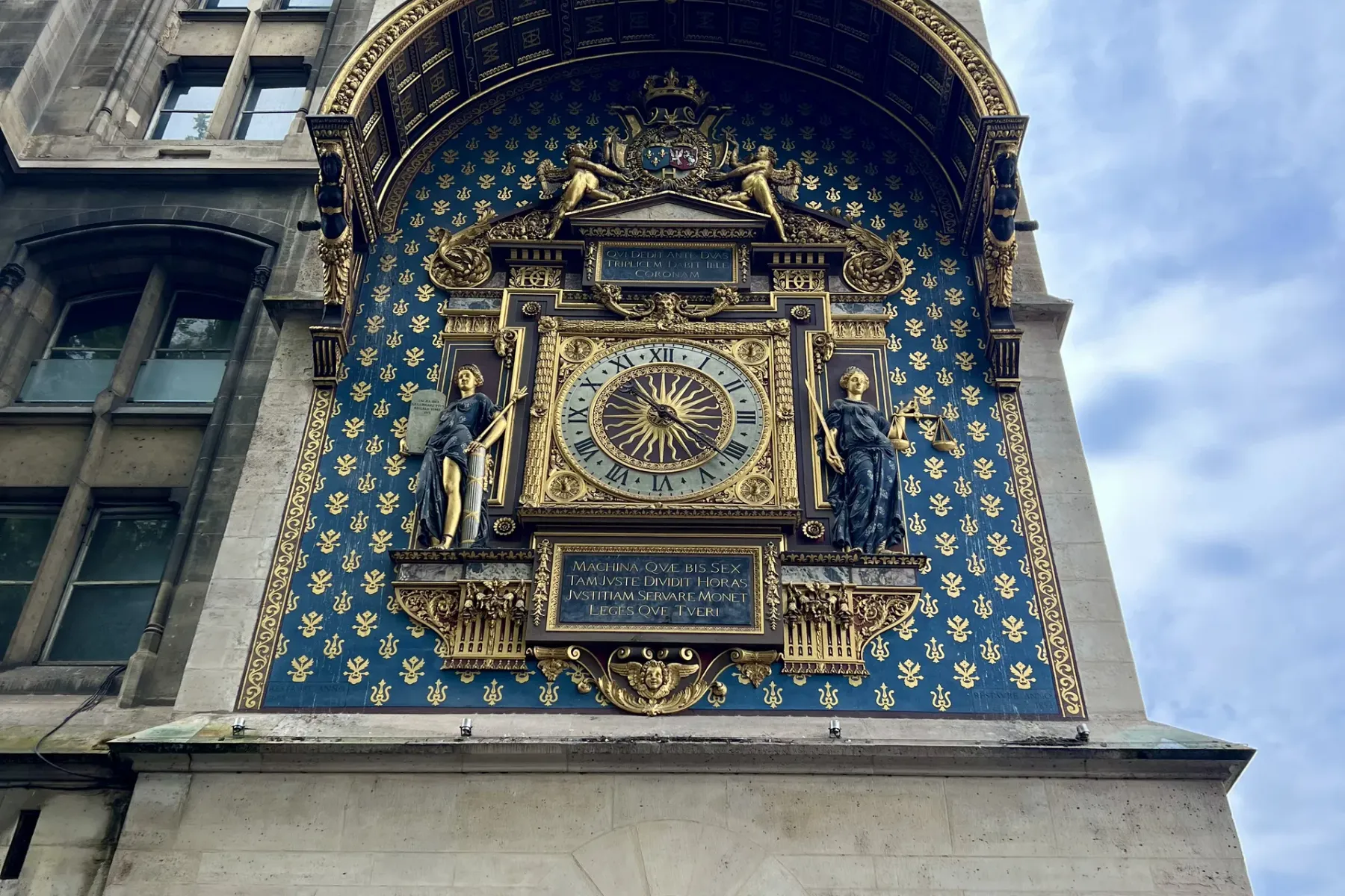 Ornate golden clock on a historical stone tower with blue and gold fleur-de-lis pattern, flanked by two statues. Paris str...