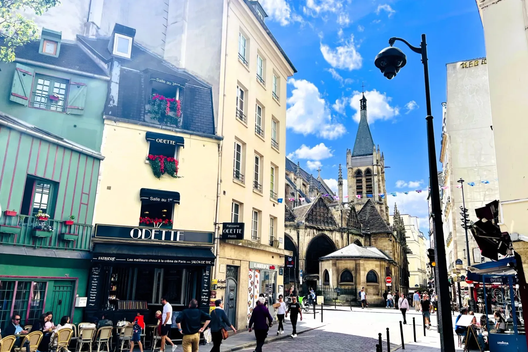 Cobblestone street in Paris with historic buildings, Odette patisserie, and Saint-Séverin church spire under a blue sky. P...