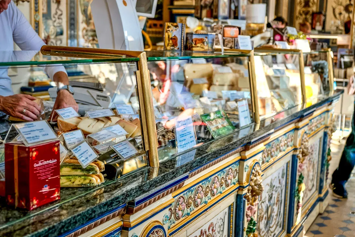 A decorated deli counter filled with various cheeses, price tags, and other products. A person's hands are visible packagi...
