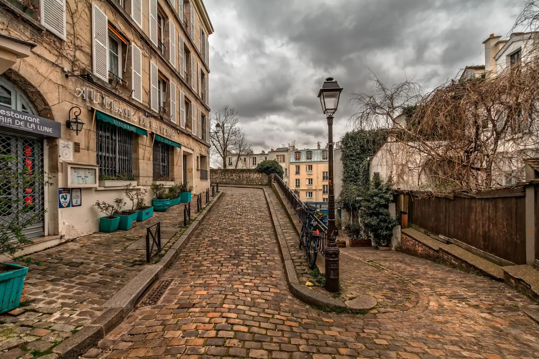 Winding cobblestone street in Paris, with a stone building on the left housing a restaurant, and a lamppost on the right.
