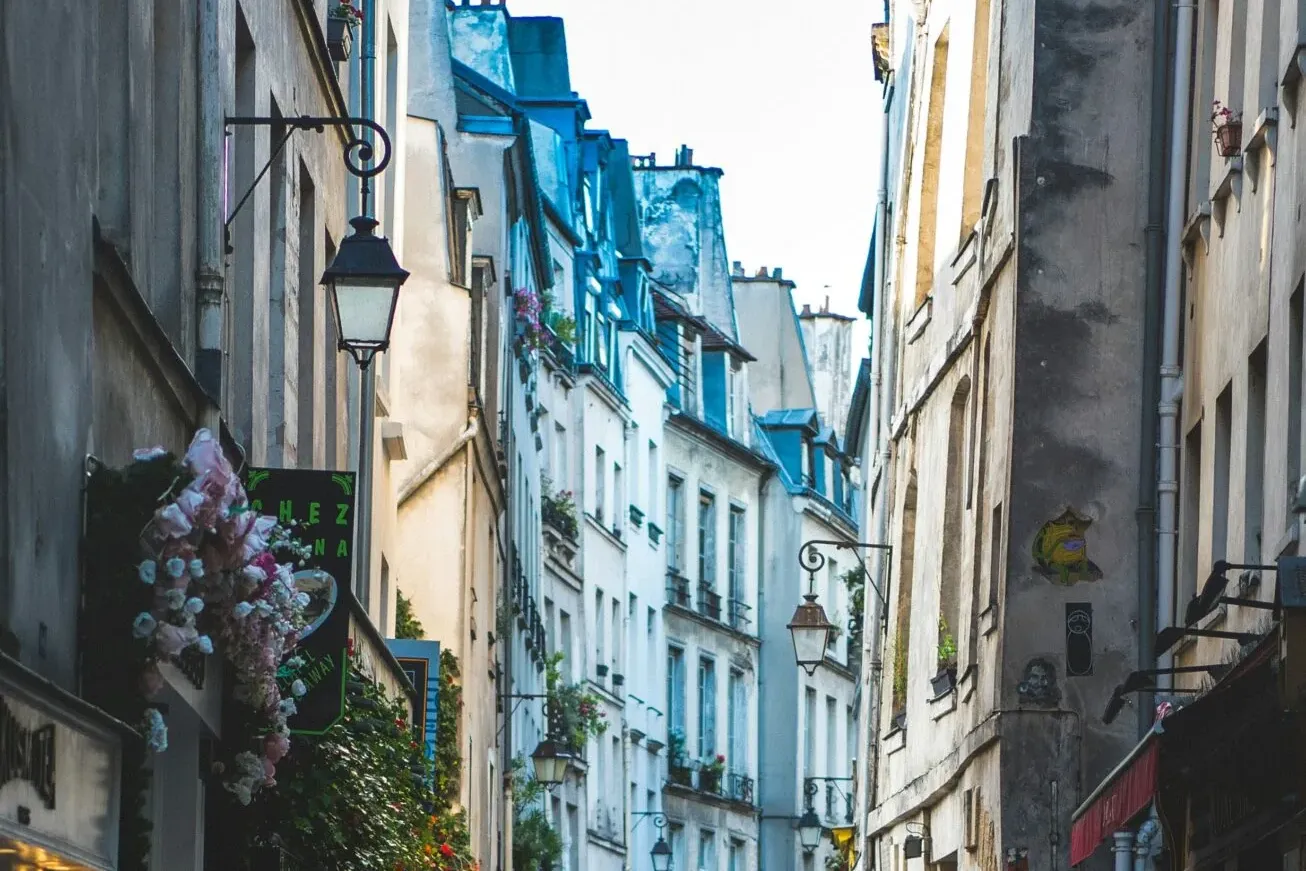 A narrow cobblestone street in Paris with old buildings, storefronts, and two people walking in the distance.