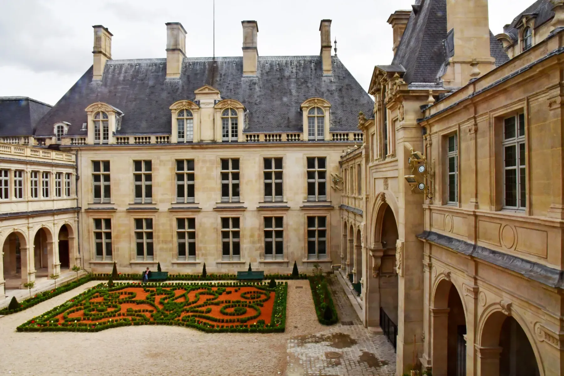 A grand Parisian courtyard with an intricately shaped formal garden, surrounded by light-colored stone buildings with dorm...