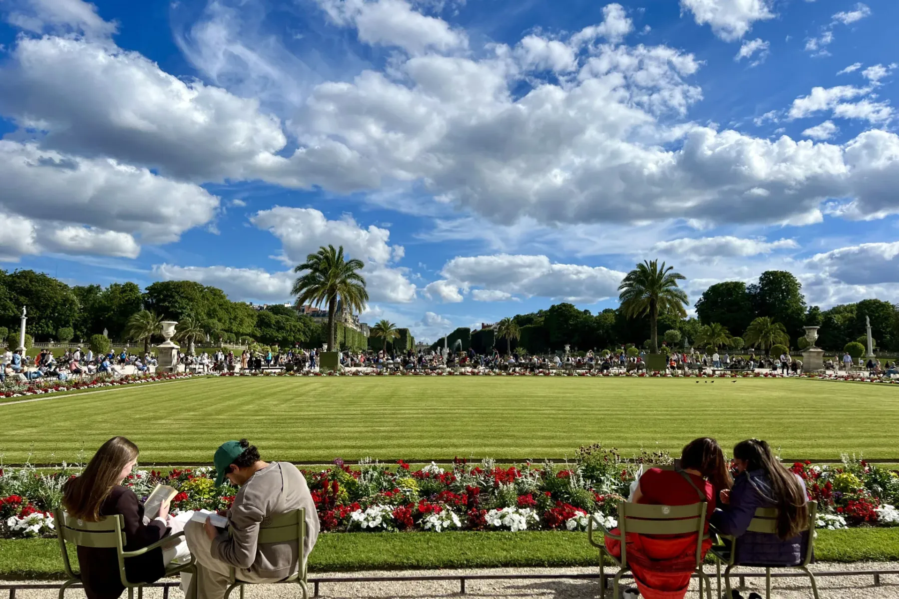 People sitting on green chairs, reading in a large formal garden with a wide lawn, flowerbeds, and palm trees under a clou...