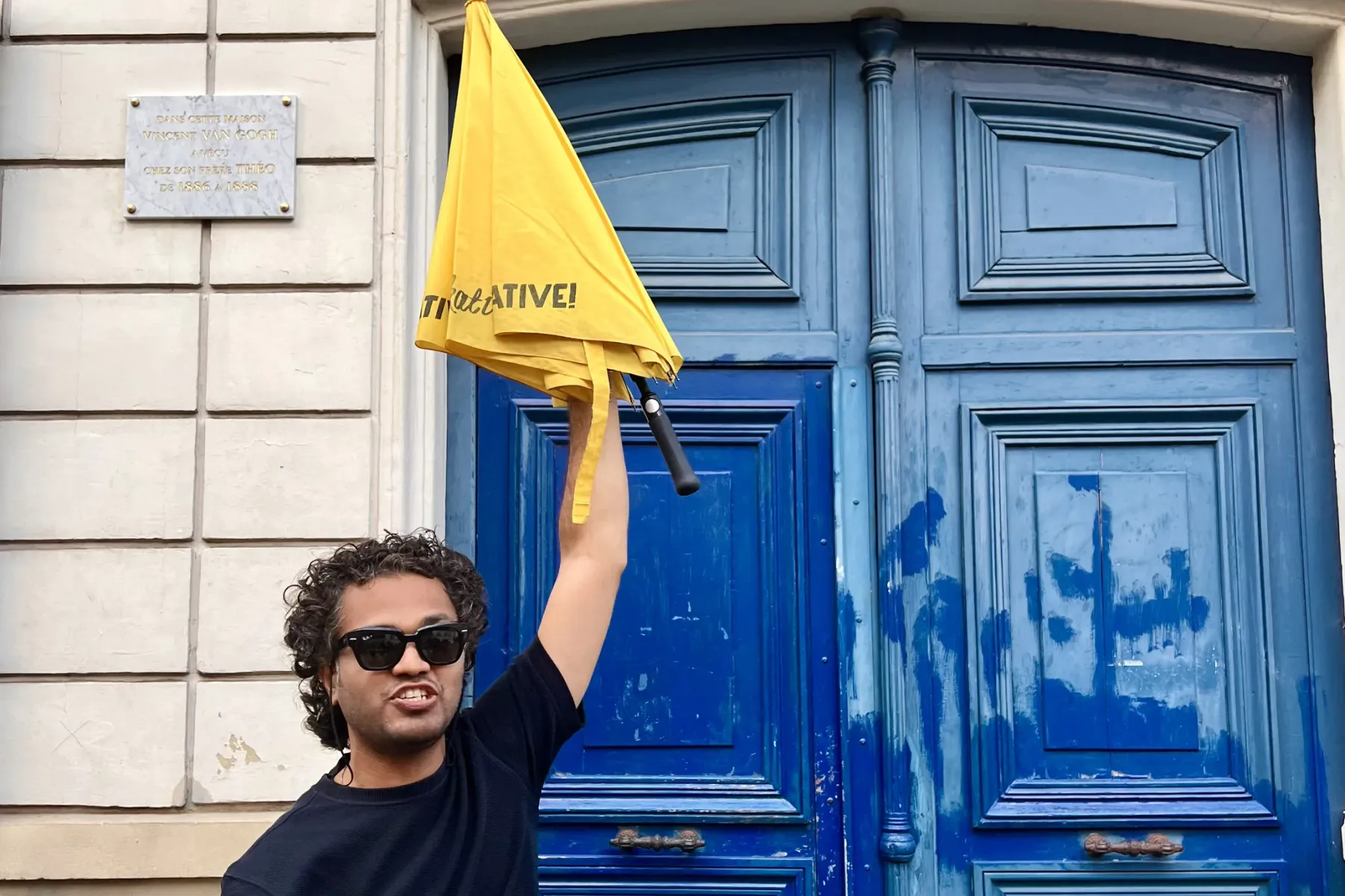 Man holding yellow Walkative! umbrella in front of a blue door with '54' and a plaque about Van Gogh's residence.