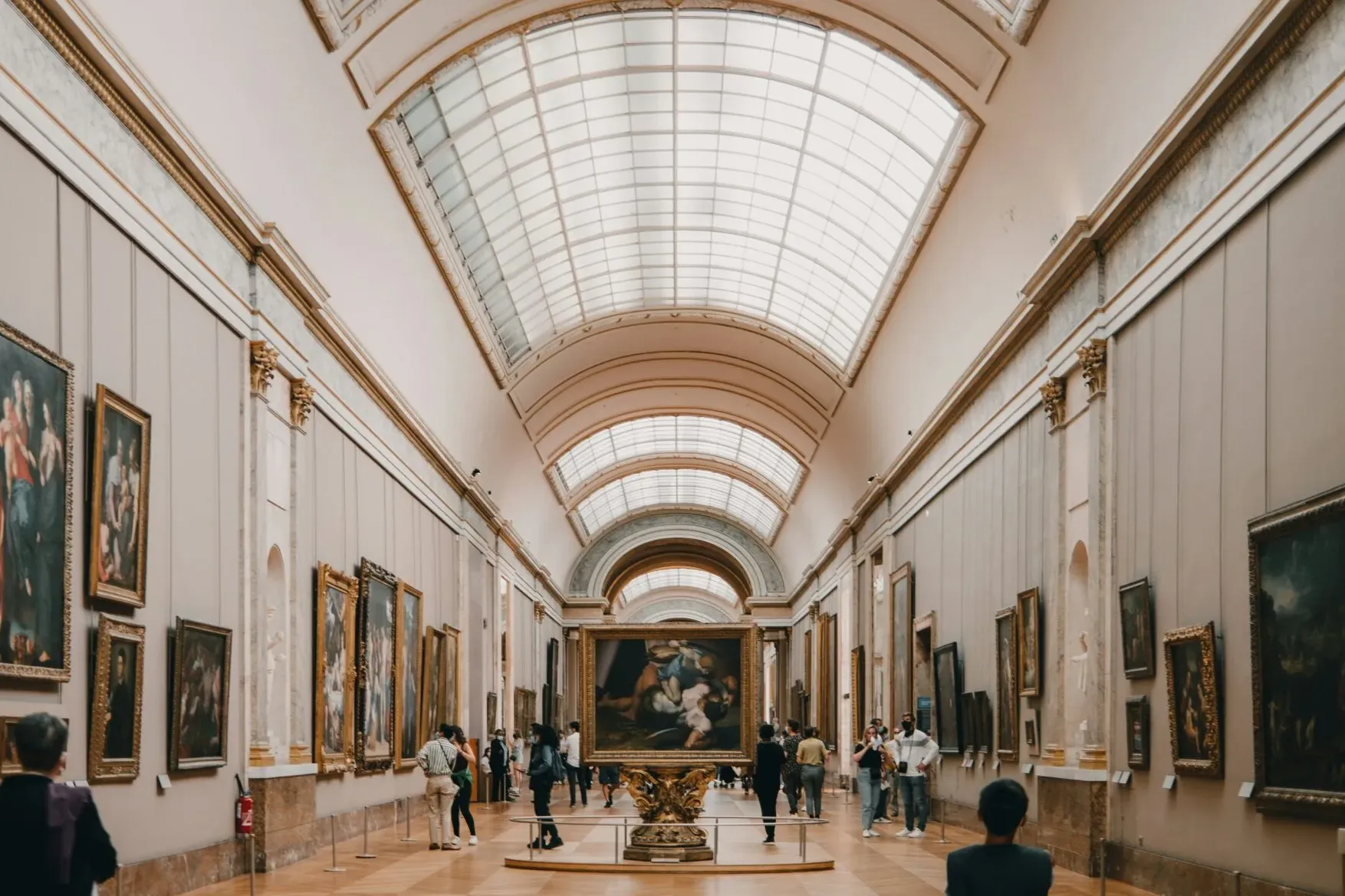 Grand art gallery in Paris with numerous classical paintings lining the walls and a large vaulted skylight ceiling. Visito...