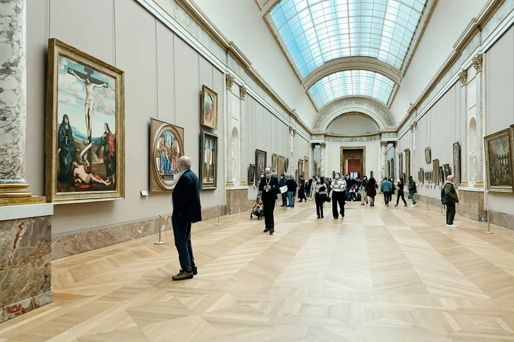 Long museum gallery with a wooden parquet floor, paintings on light walls, and several visitors. A large skylight illumina...