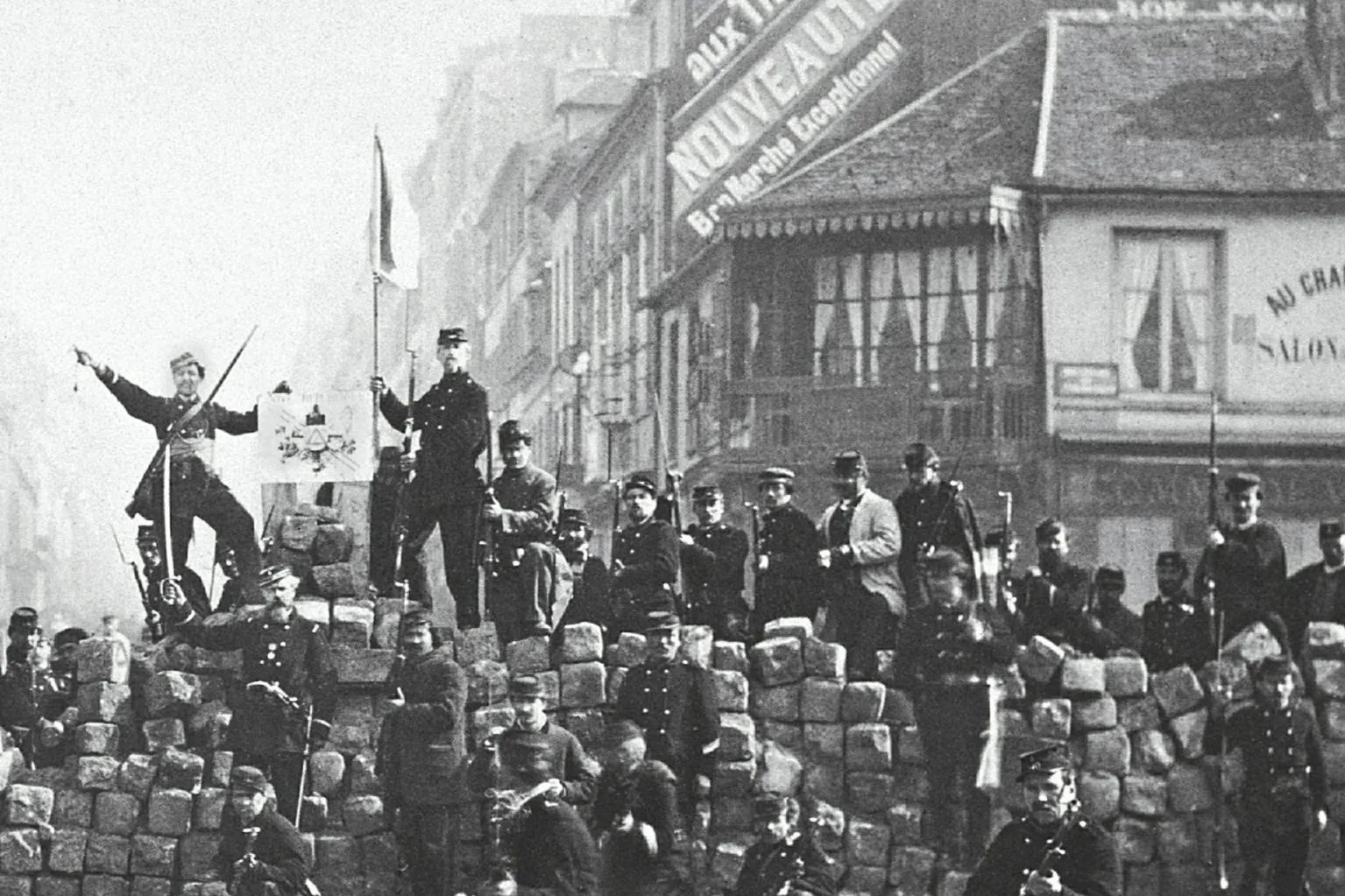 Black and white photo shows men in military uniforms on a cobblestone barricade in a Paris street with historic buildings.